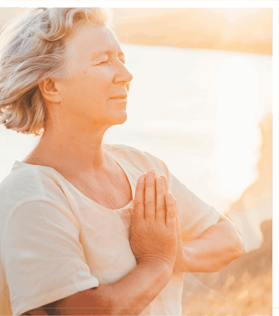 mature blonde woman standing in front of a body of water at sunrise with her hands in prayer position and eyes closed